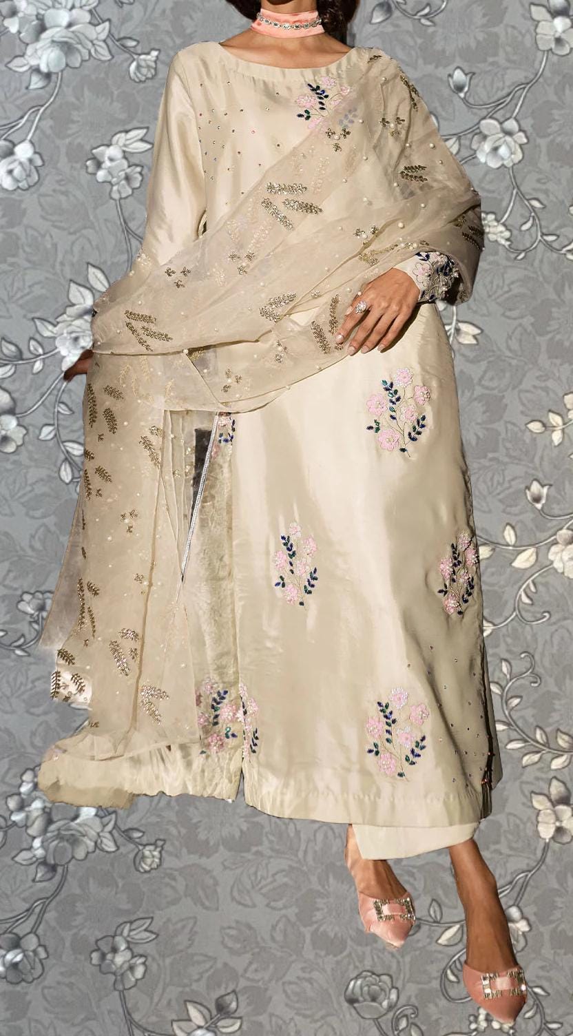A woman wearing a beige dress with floral embroidery and a matching shawl stands against a gray floral background.