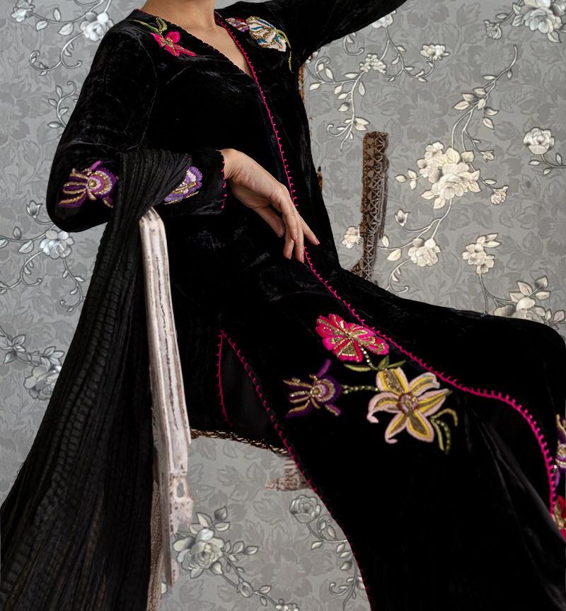 A person wearing a long black dress with colorful floral embroidery, sitting against a gray floral background.