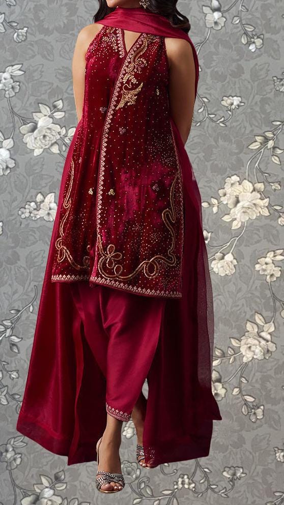 A woman wearing a red and gold embroidered dress with a high neckline and a long skirt, standing against a gray floral background.