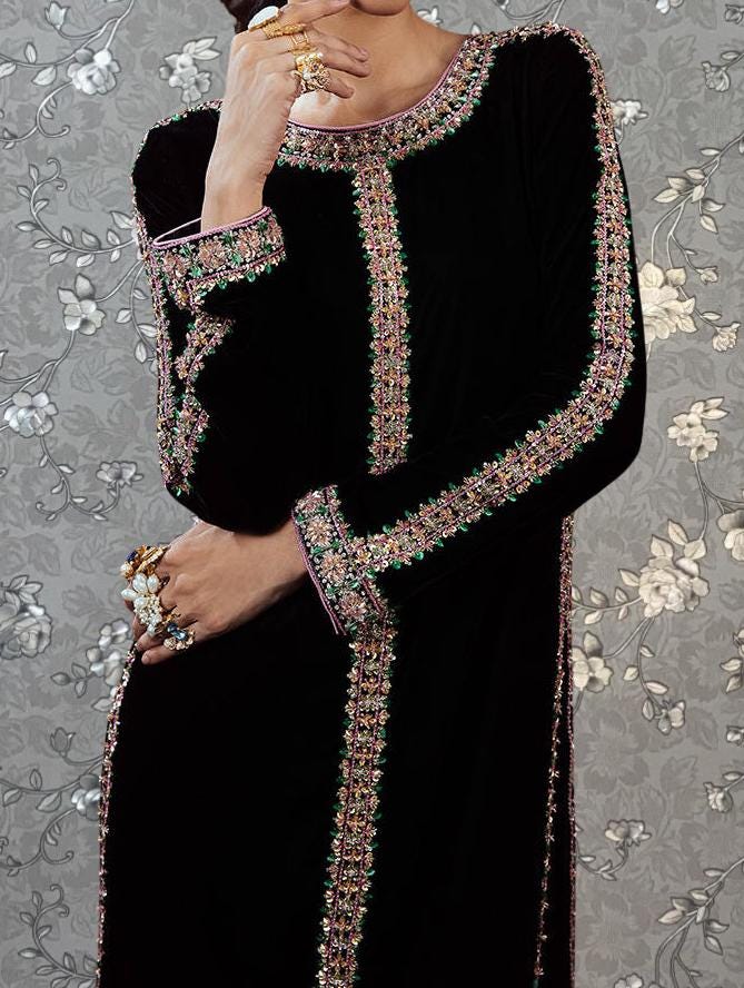 A woman wearing a long black dress with intricate embroidery and jewelry poses against a floral background.