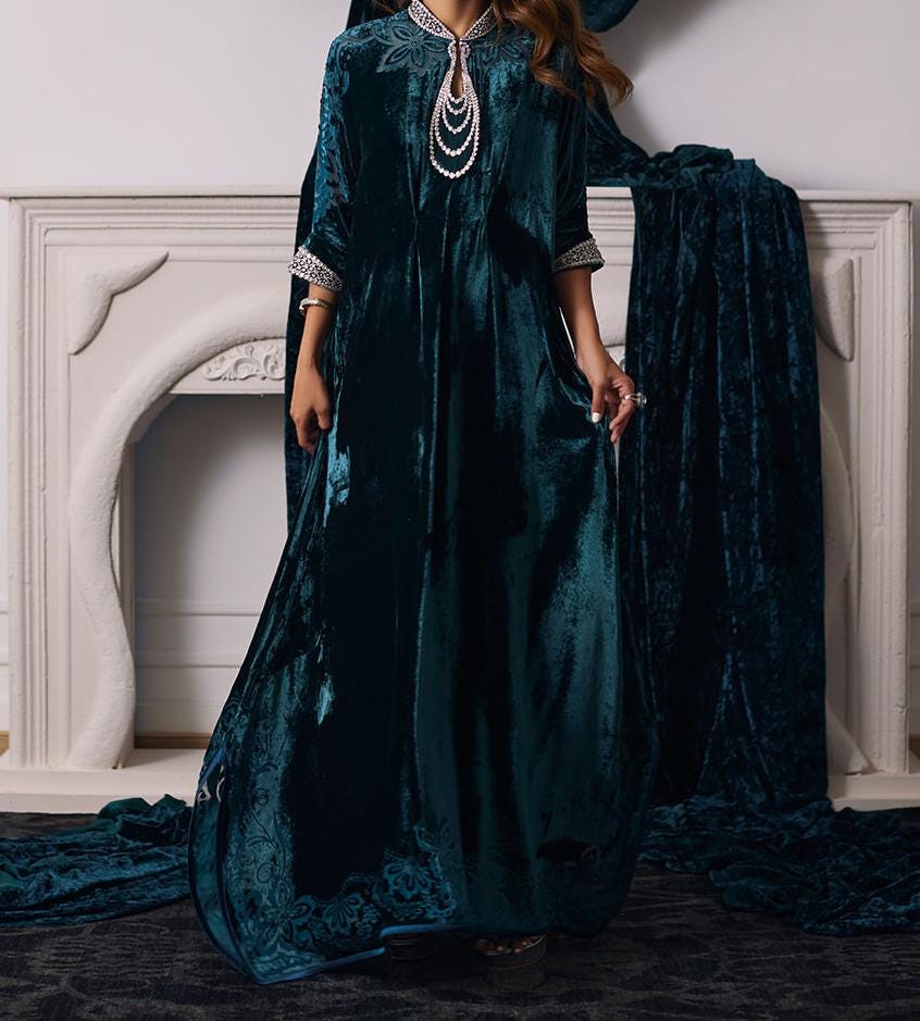 A woman wearing a long, dark blue velvet dress with a high neckline and a large, ornate necklace stands in front of a white fireplace.