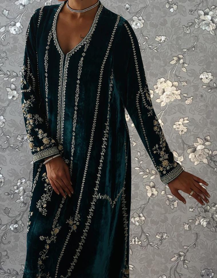A person wearing a long, dark green velvet dress with intricate gold embroidery stands against a gray floral background.