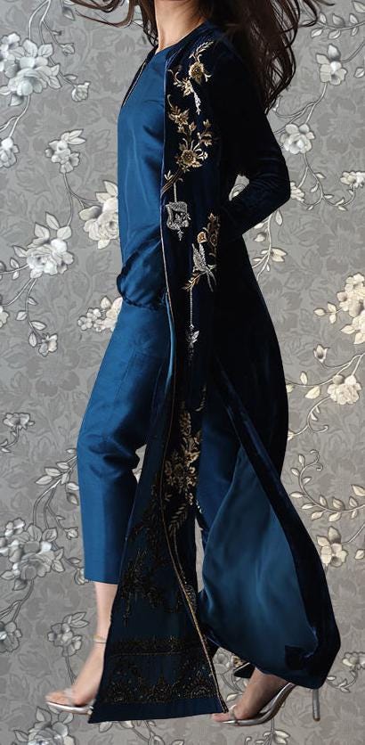 A woman wearing a long blue dress with gold embroidery and a matching long blue coat, standing against a gray floral background.