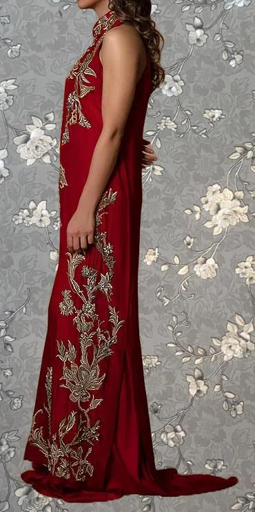 A woman wearing a long, red dress with intricate gold embroidery stands against a gray floral background.