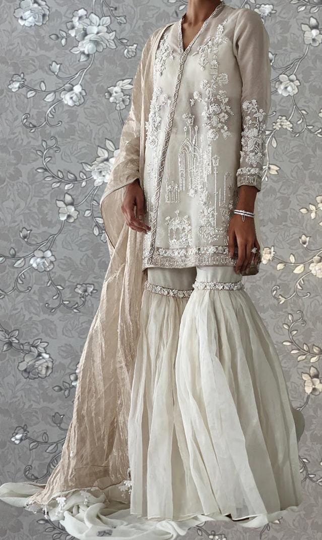 A woman wearing a long, white dress with intricate embroidery and a matching skirt, standing against a gray floral background.