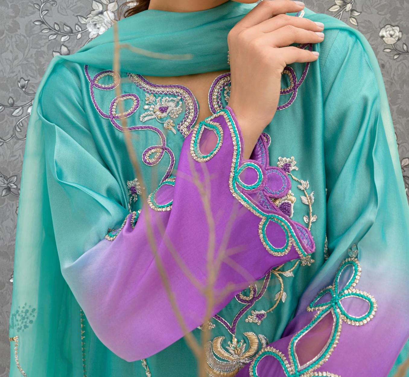 A close-up of a person wearing a turquoise and purple dress with intricate embroidery, posing with their hand on their hip.