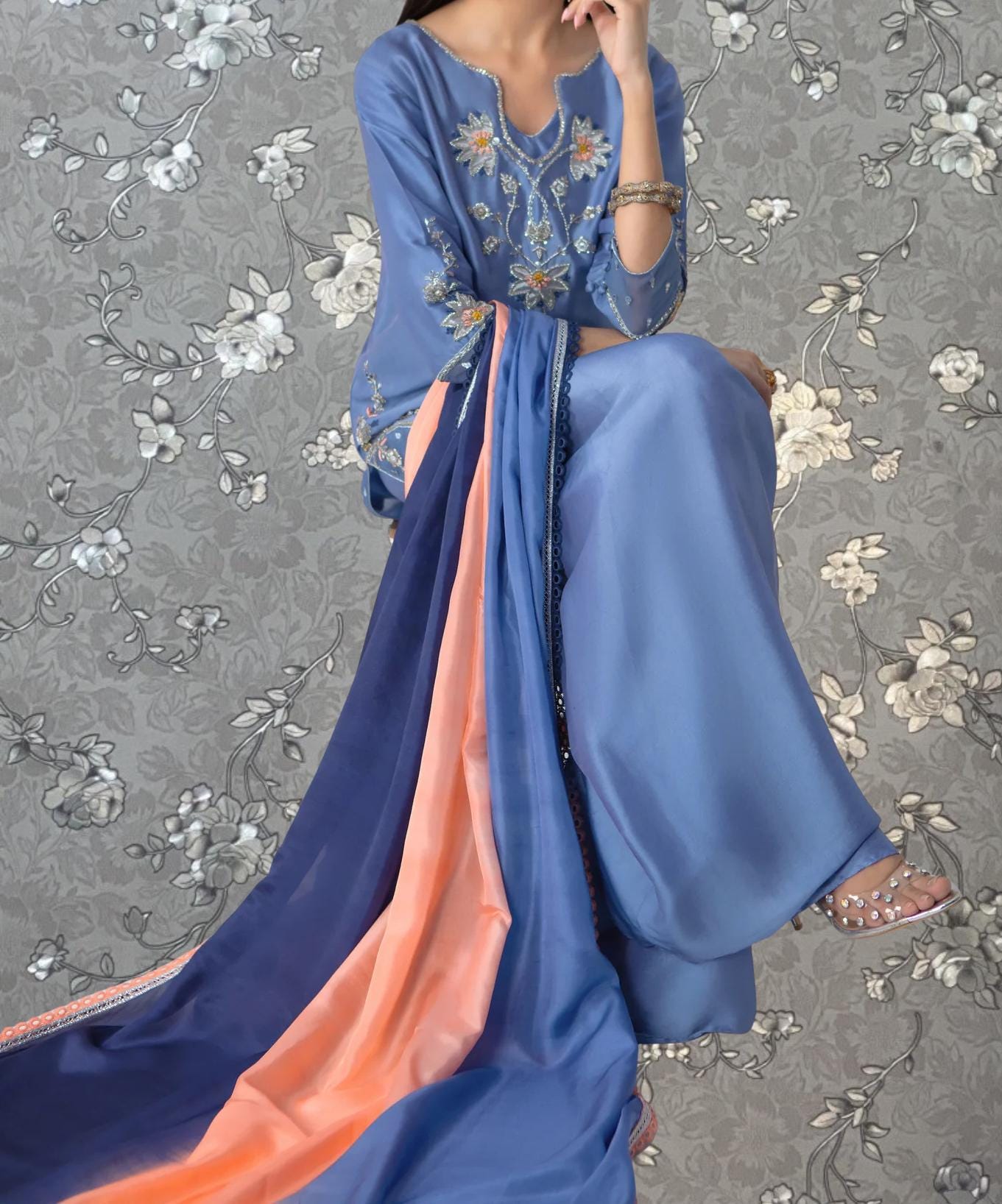 A woman wearing a blue dress with a long, flowing skirt and a matching blue and orange shawl is sitting against a gray floral background.