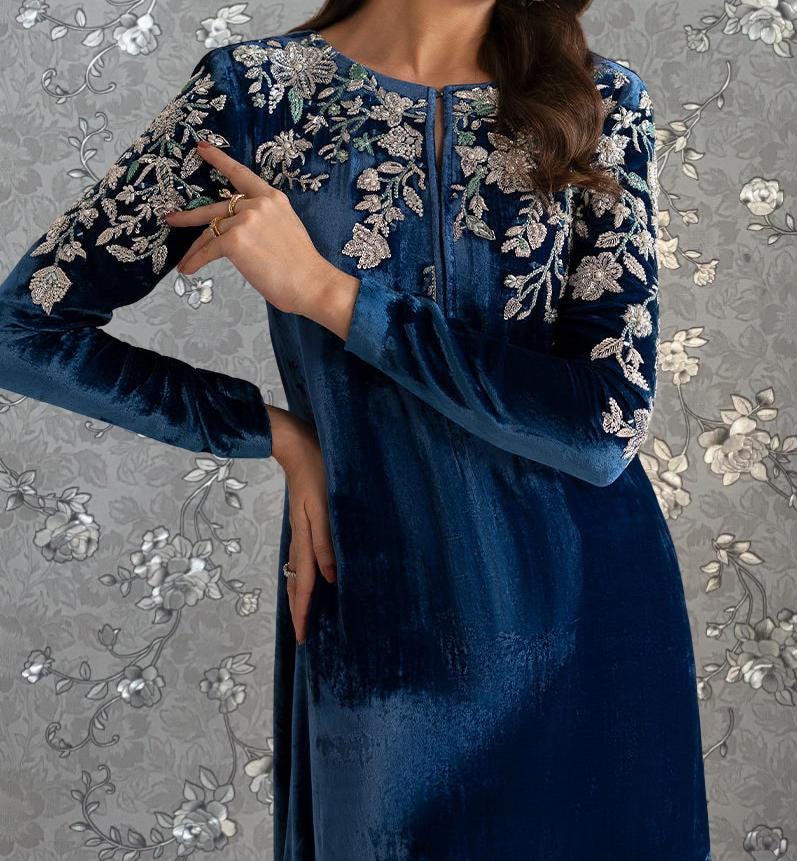 A woman wearing a long, blue velvet dress with intricate floral embroidery on the sleeves and neckline.