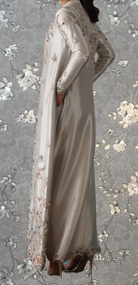 A person wearing a long, flowing, beige dress with intricate floral embroidery stands against a gray floral background.