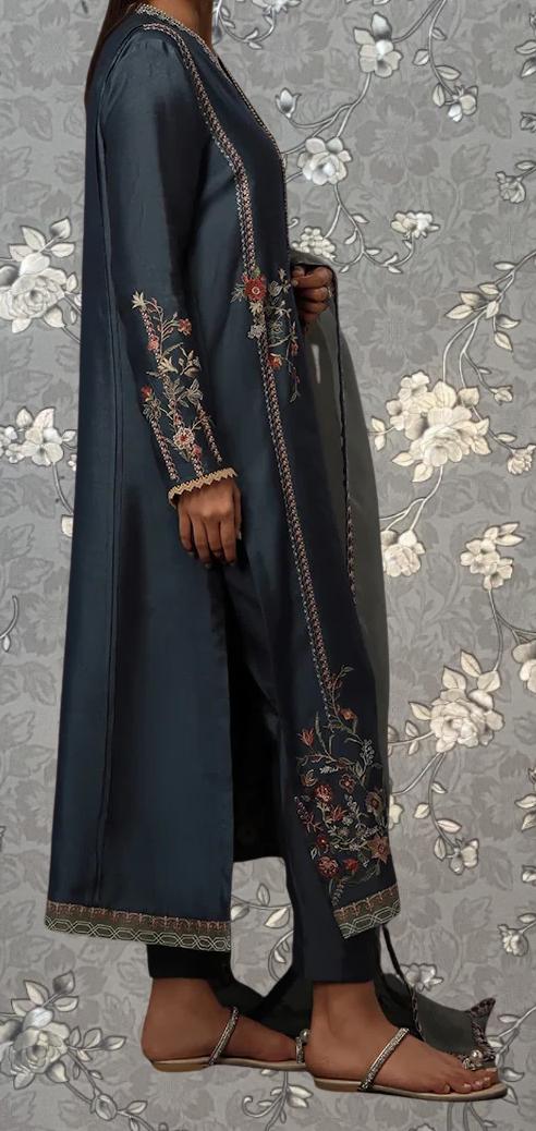 A person wearing a long, dark blue dress with intricate floral embroidery stands against a gray floral background.