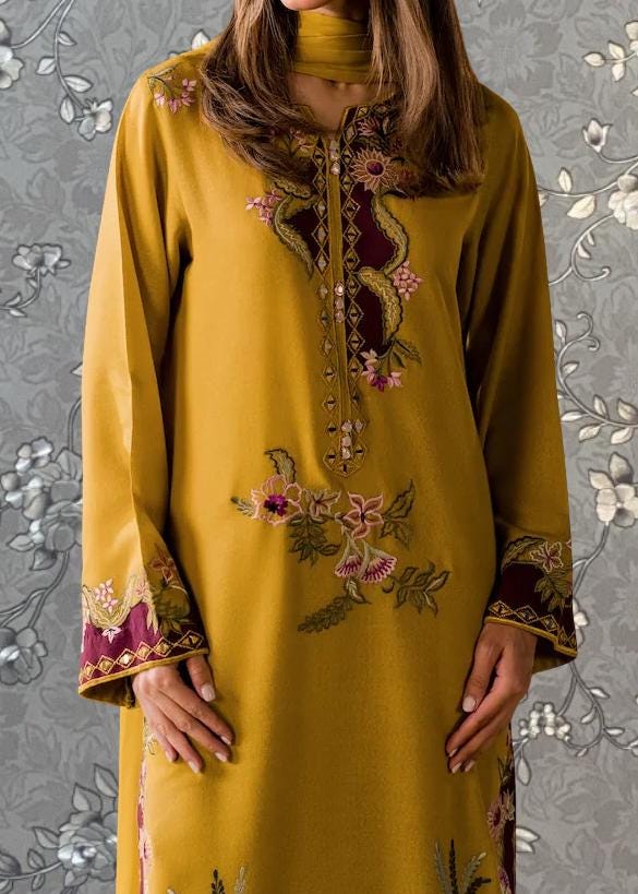 A woman wearing a yellow dress with intricate floral embroidery stands against a gray floral background.