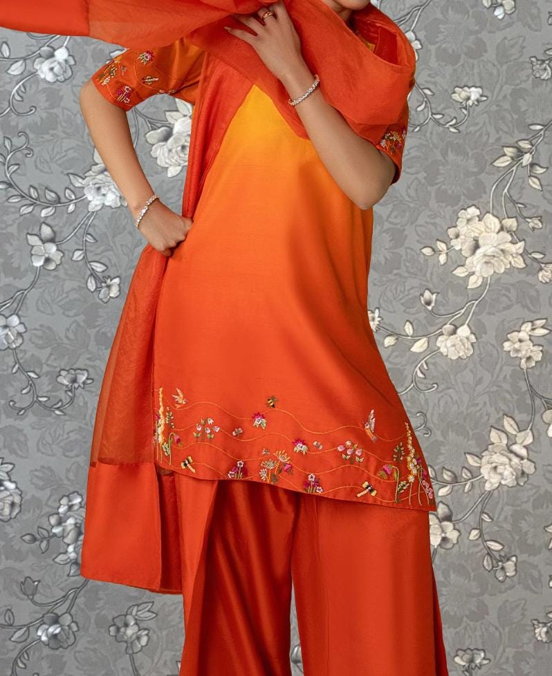 A person wearing a vibrant orange dress with floral embroidery stands against a gray floral background.