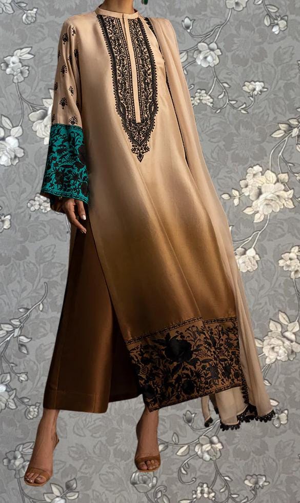 A woman wearing a long, beige dress with intricate black embroidery and floral patterns, standing against a gray floral background.