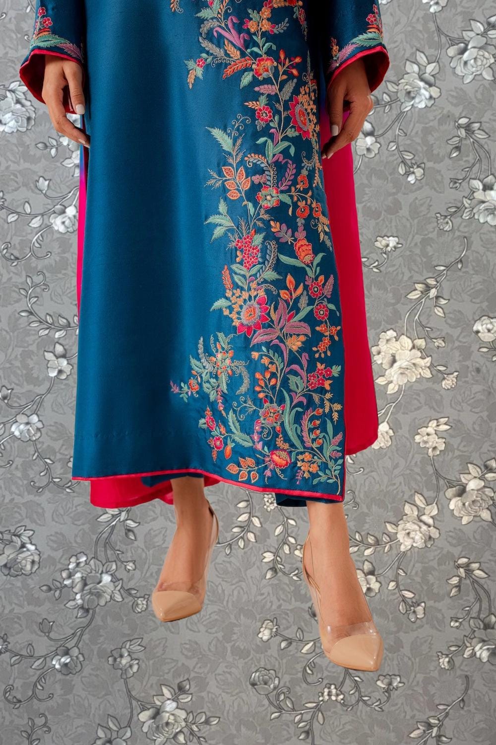 A person wearing a long, blue dress with a floral pattern and red accents, standing against a gray floral background.