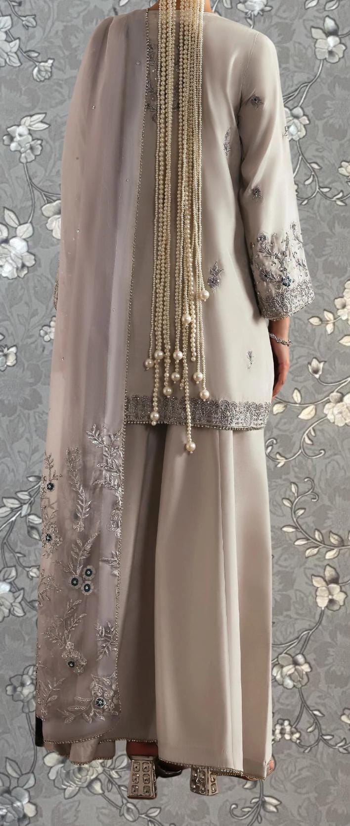 a person wearing a long, beige dress with intricate embroidery and a long, beige shawl. The dress features a high collar and long sleeves, and the person is wearing a pair of high-heeled shoes. The background is a gray floral pattern.