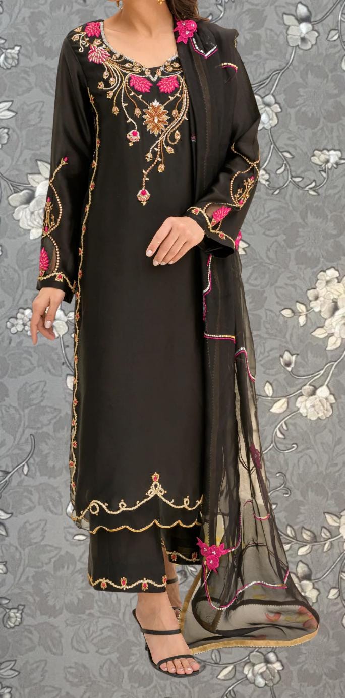 A woman wearing a black dress with intricate embroidery and pink accents, standing against a gray floral background.
