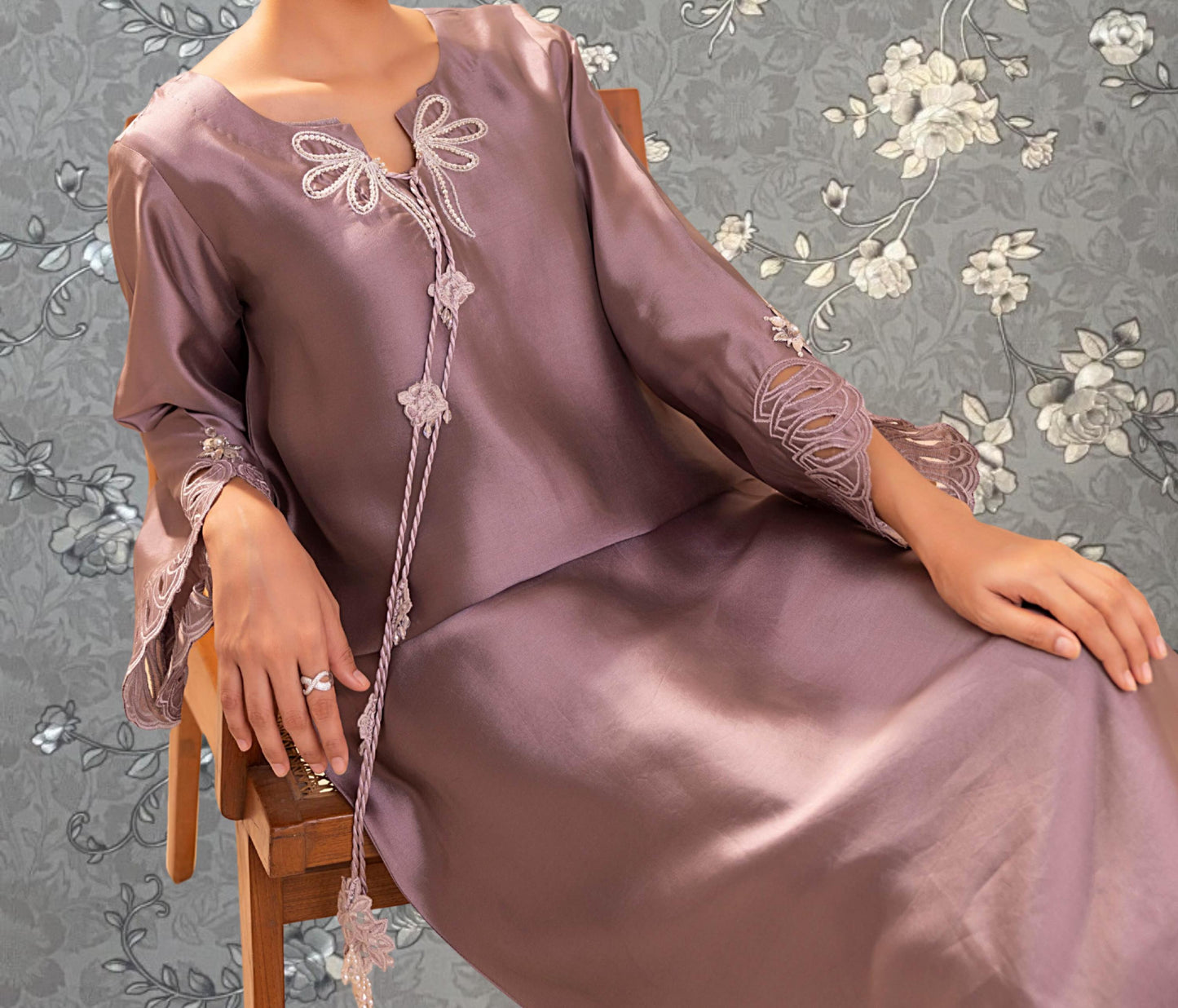 A woman wearing a purple dress with lace details and a floral pattern is sitting on a wooden chair against a gray floral background.