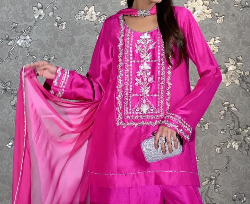 A woman wearing a bright pink dress with intricate embroidery and a matching pink shawl, holding a small silver clutch purse.