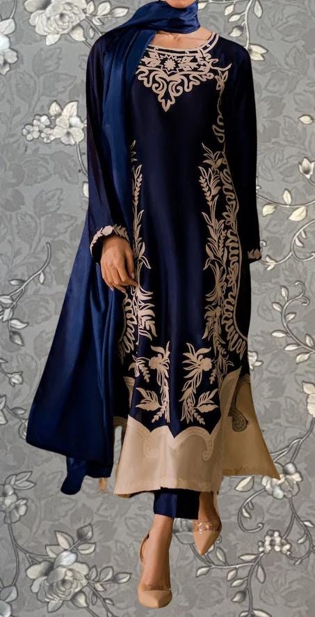 A woman wearing a blue and gold dress with intricate embroidery, standing against a gray floral background.