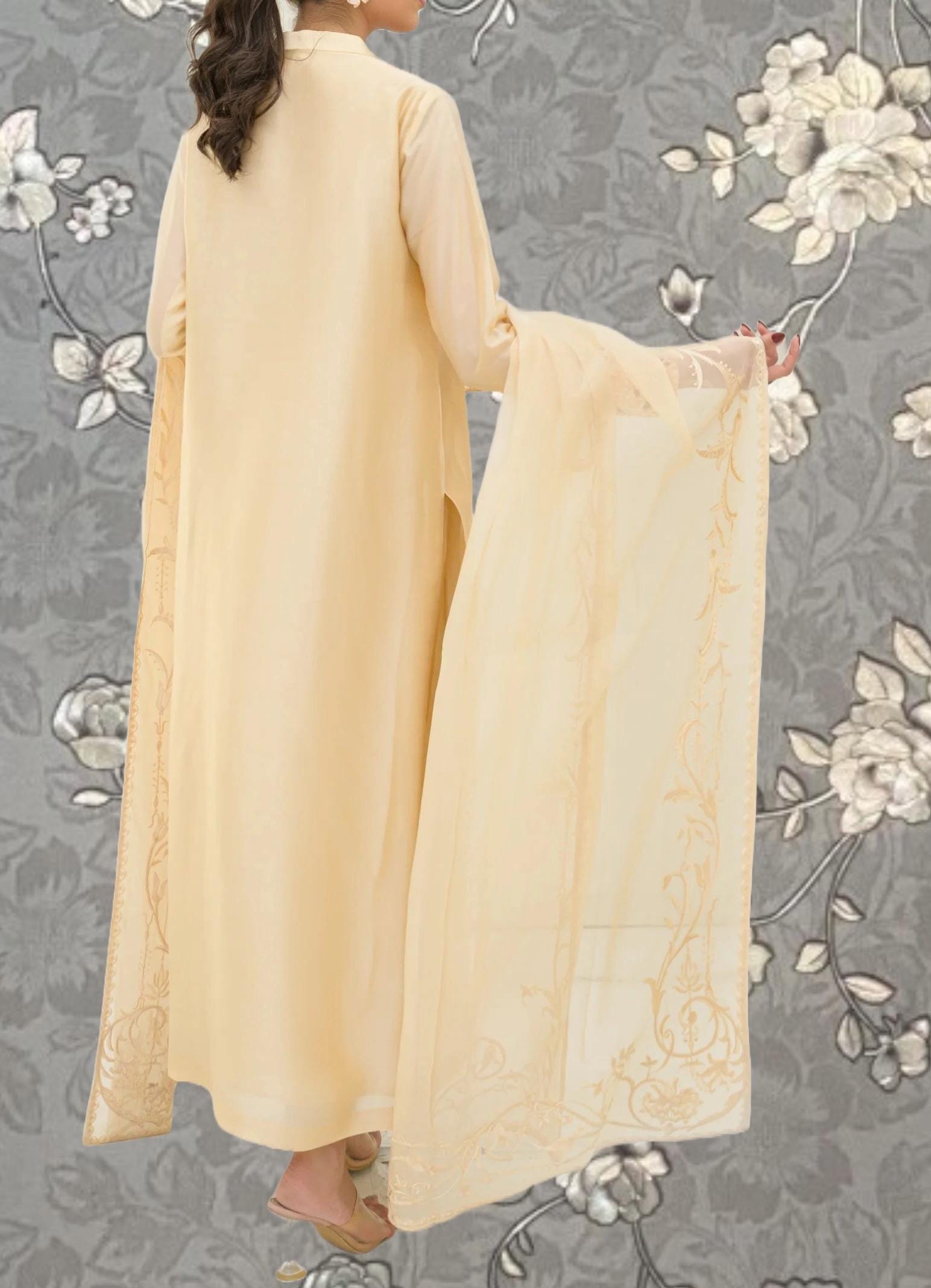 A woman wearing a long, cream-colored dress with a matching shawl stands against a gray floral background.