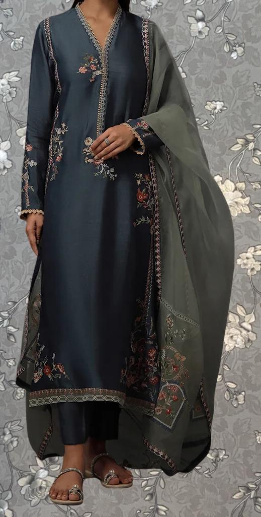 A woman wearing a long, dark blue dress with intricate embroidered patterns on the front and sides. She is wearing a matching green shawl or shawl-like garment, and her feet are adorned with silver sandals. The background features a floral pattern, which complements the overall design of the outfit.