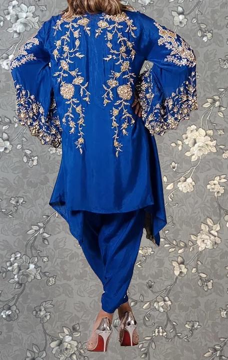 A person wearing a blue dress with gold embroidery and a matching blue jacket with gold embroidery, standing against a gray floral background.
