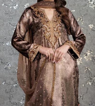 A woman wearing a brown and gold dress with intricate embroidery and a matching shawl stands against a gray floral background.