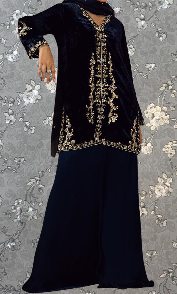 A person wearing a long black dress with gold embroidery stands against a gray floral background.