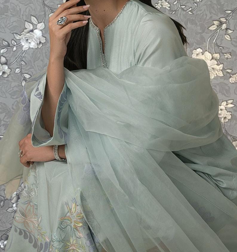 A woman wearing a light blue dress with a sheer, flowing fabric draped over her. She is holding her hand to her face, and there is a floral pattern on the background.