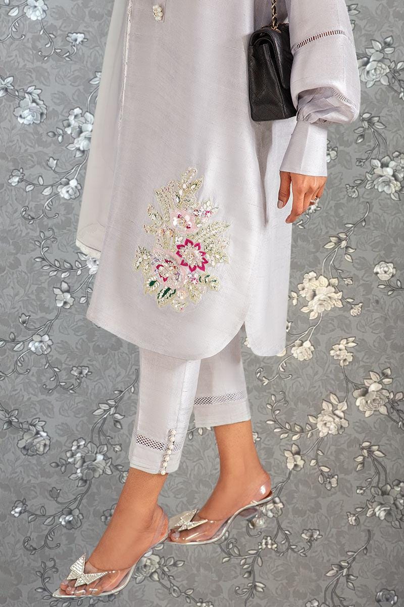 A person wearing a white dress with a floral embroidered design, standing against a gray floral background.