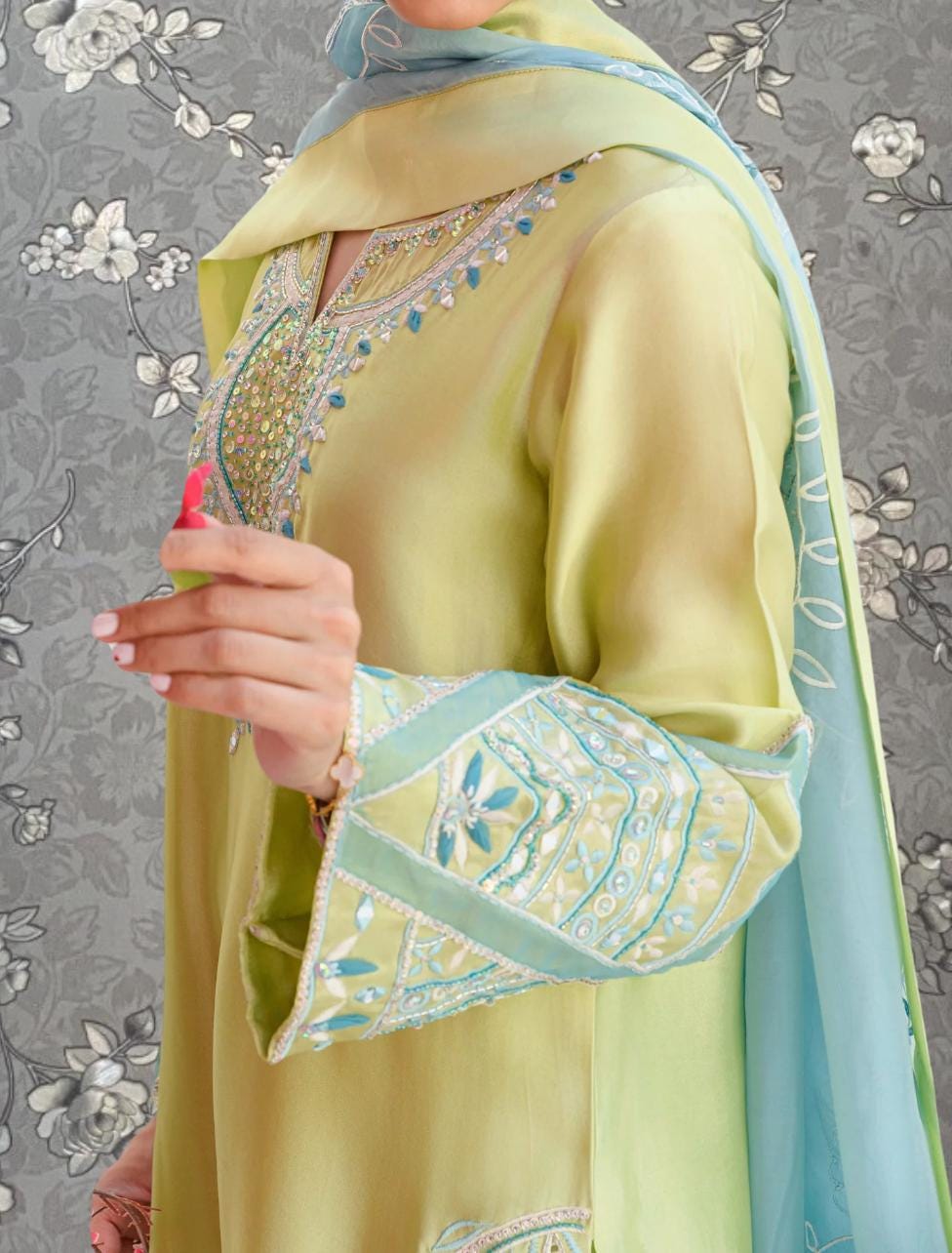 A person wearing a yellow and green dress with intricate embroidery and a matching scarf stands against a gray floral background.