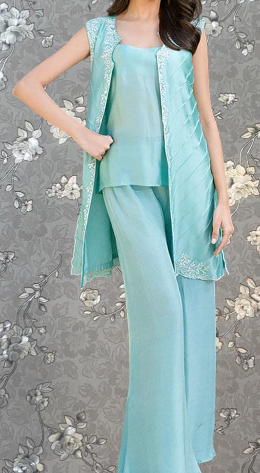 A woman wearing a light blue dress with a sleeveless design and a matching vest, standing against a gray floral background.