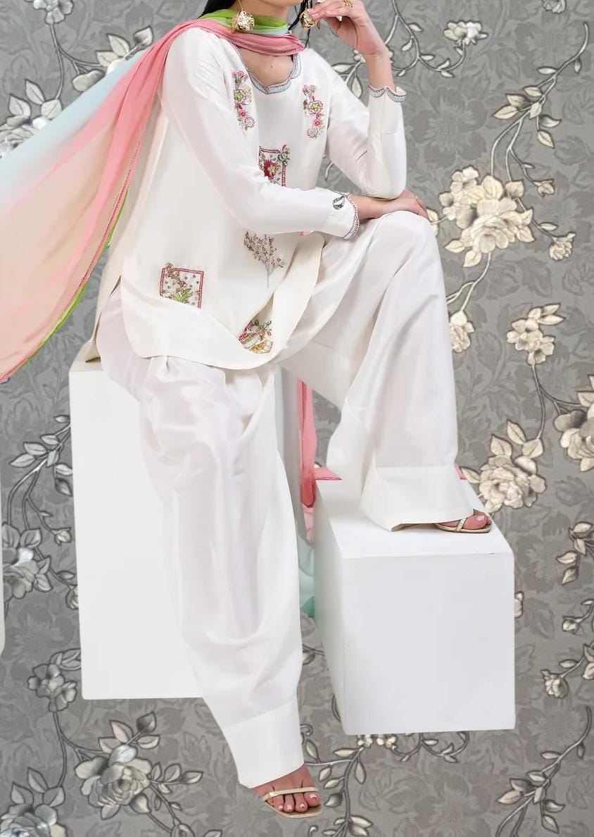 A person wearing a white outfit with floral embroidery is sitting on a white platform against a gray floral background.