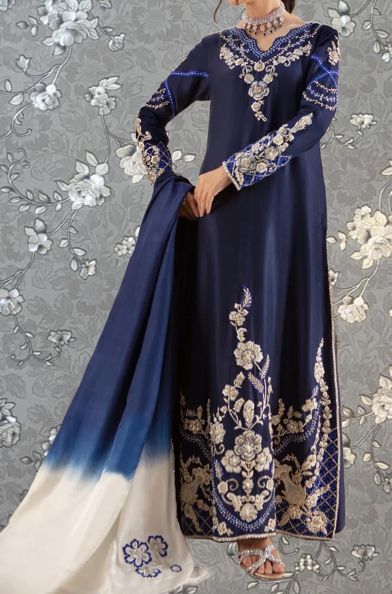 A woman wearing a long, blue dress with intricate embroidery and floral patterns, posing against a gray floral background.