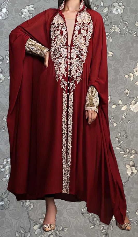 A woman wearing a long, burgundy dress with intricate embroidery and gold accents, posing against a gray floral background.