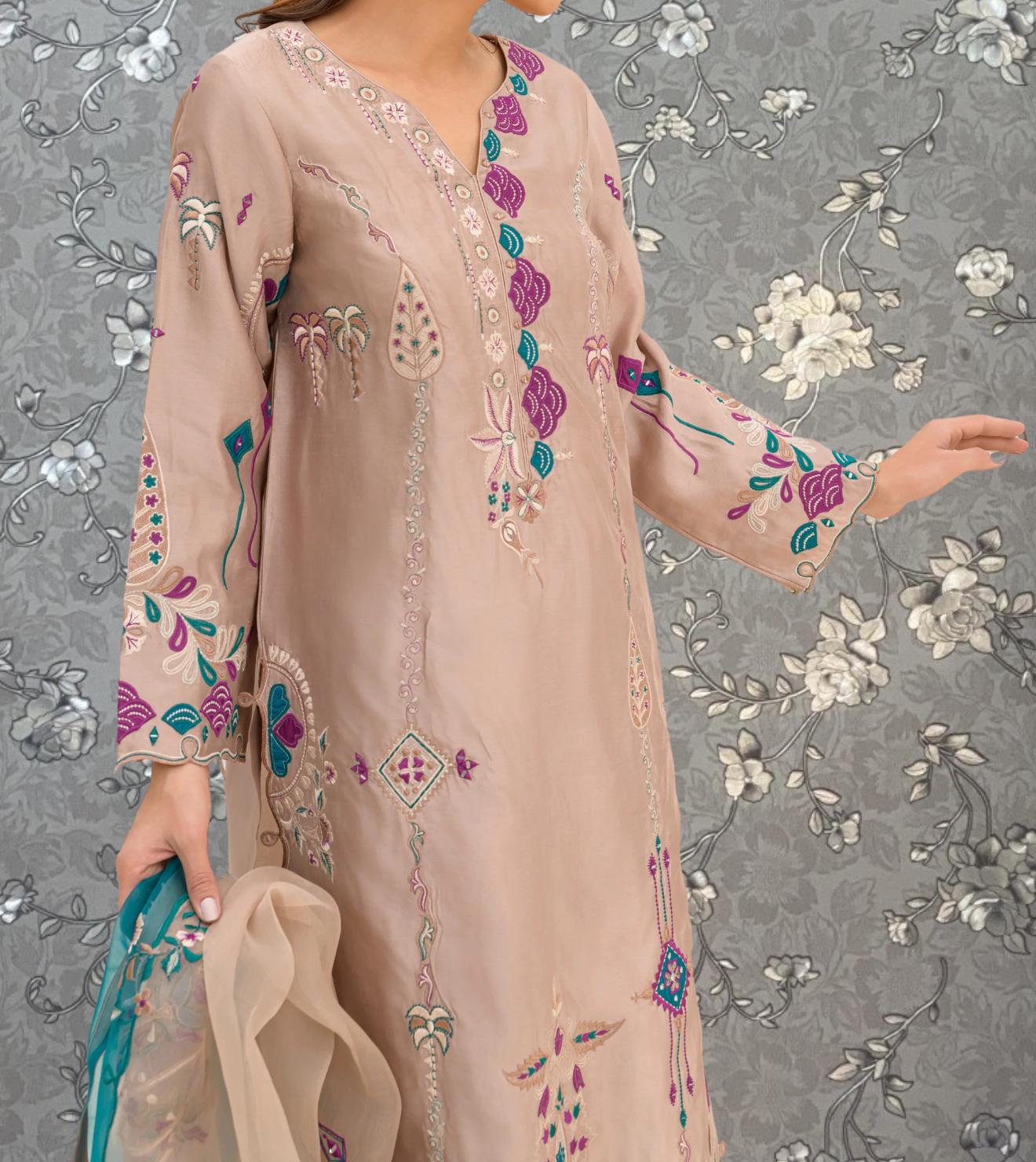 A woman wearing a long, beige dress with intricate floral embroidery stands against a gray floral background.