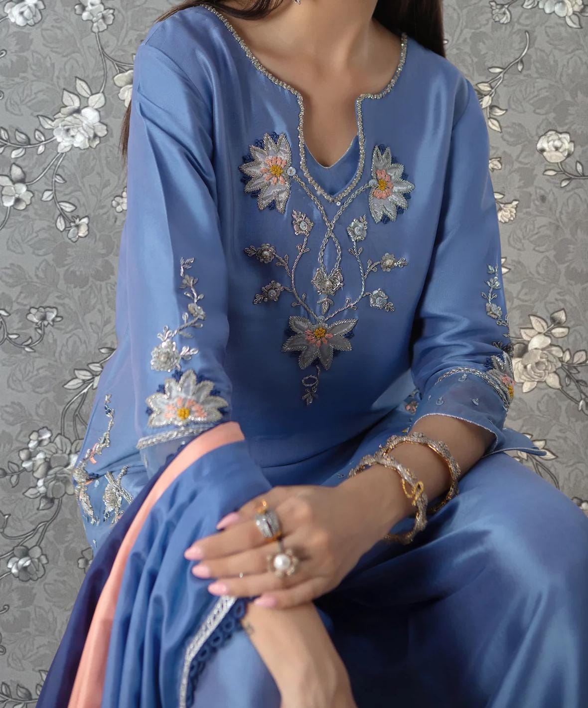 A woman wearing a blue dress with intricate embroidered designs, sitting against a gray floral background.