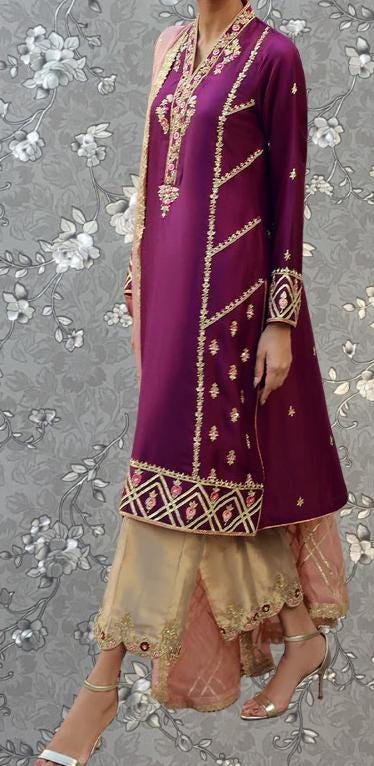 A woman wearing a long purple dress with gold embroidery and a matching skirt, standing against a gray floral background.