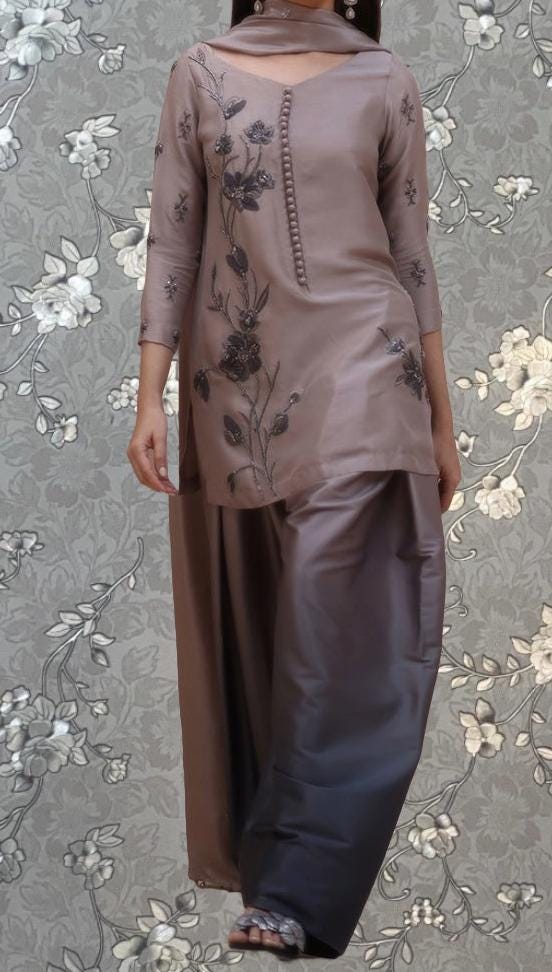 A woman wearing a brown dress with floral embroidery and a matching skirt, standing against a gray floral background.