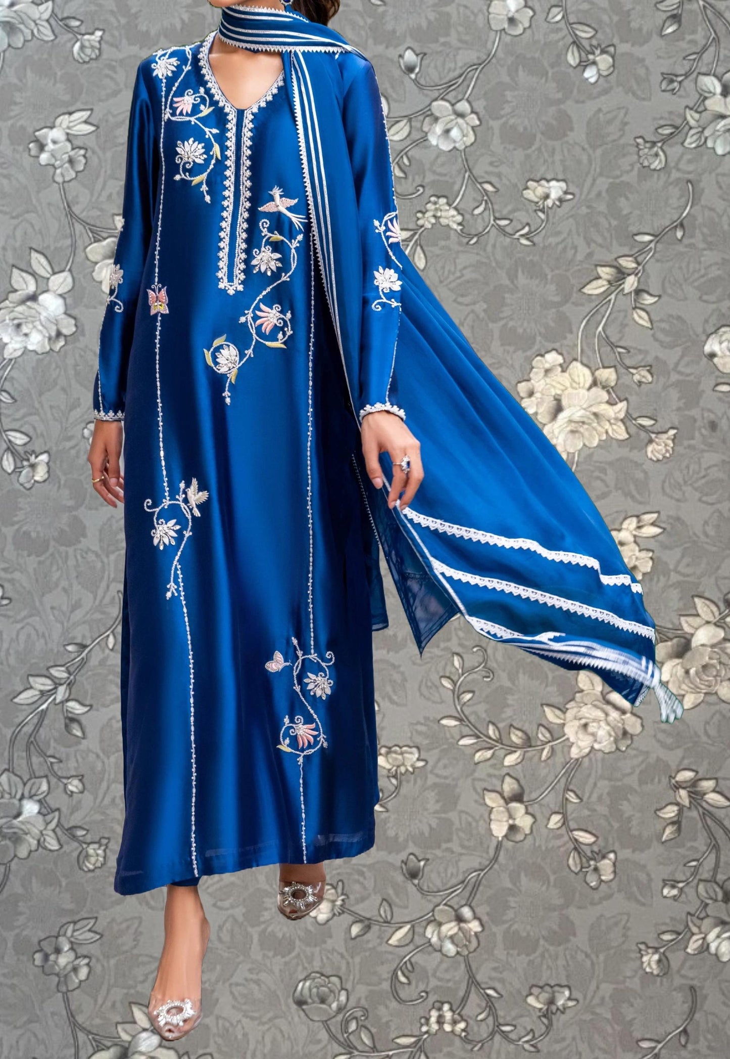 A woman wearing a long, blue dress with intricate floral embroidery stands against a gray floral background.