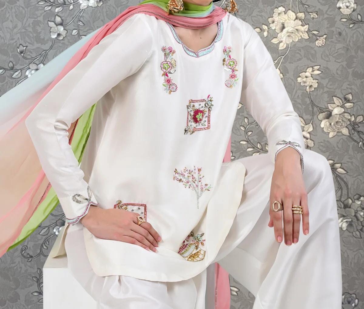 A person wearing a white dress with floral embroidery and a pink scarf is posing against a gray floral background.