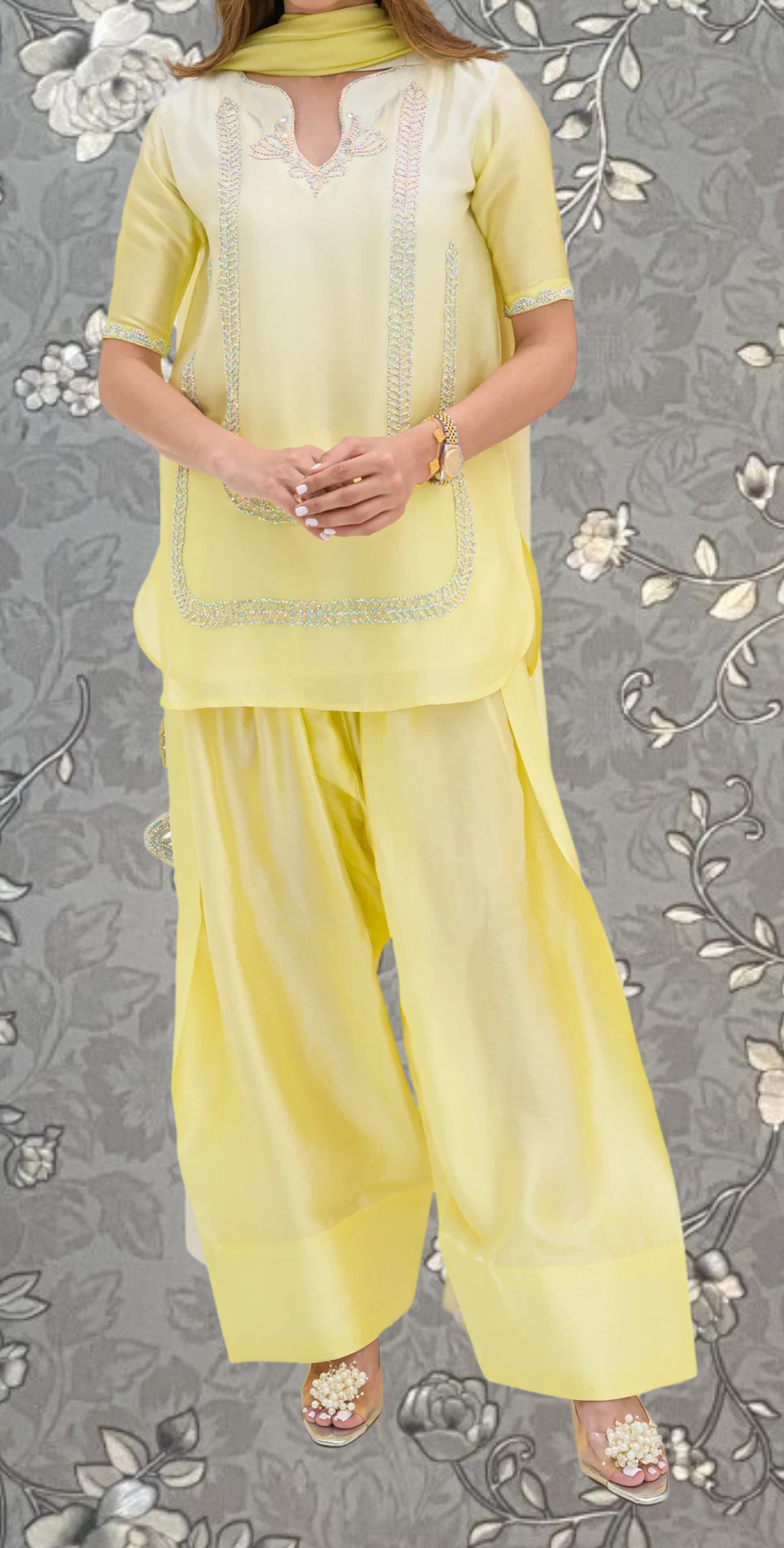 A woman wearing a yellow dress with a floral pattern and matching yellow pants stands against a gray floral background.