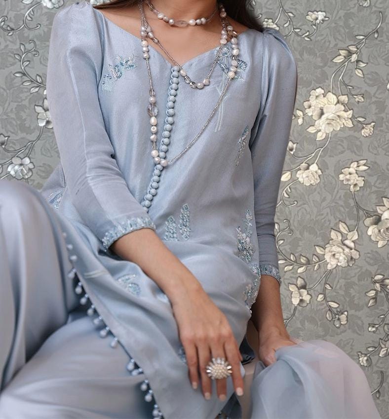 A woman wearing a light blue dress with floral embroidery and multiple necklaces, posing against a floral patterned background.