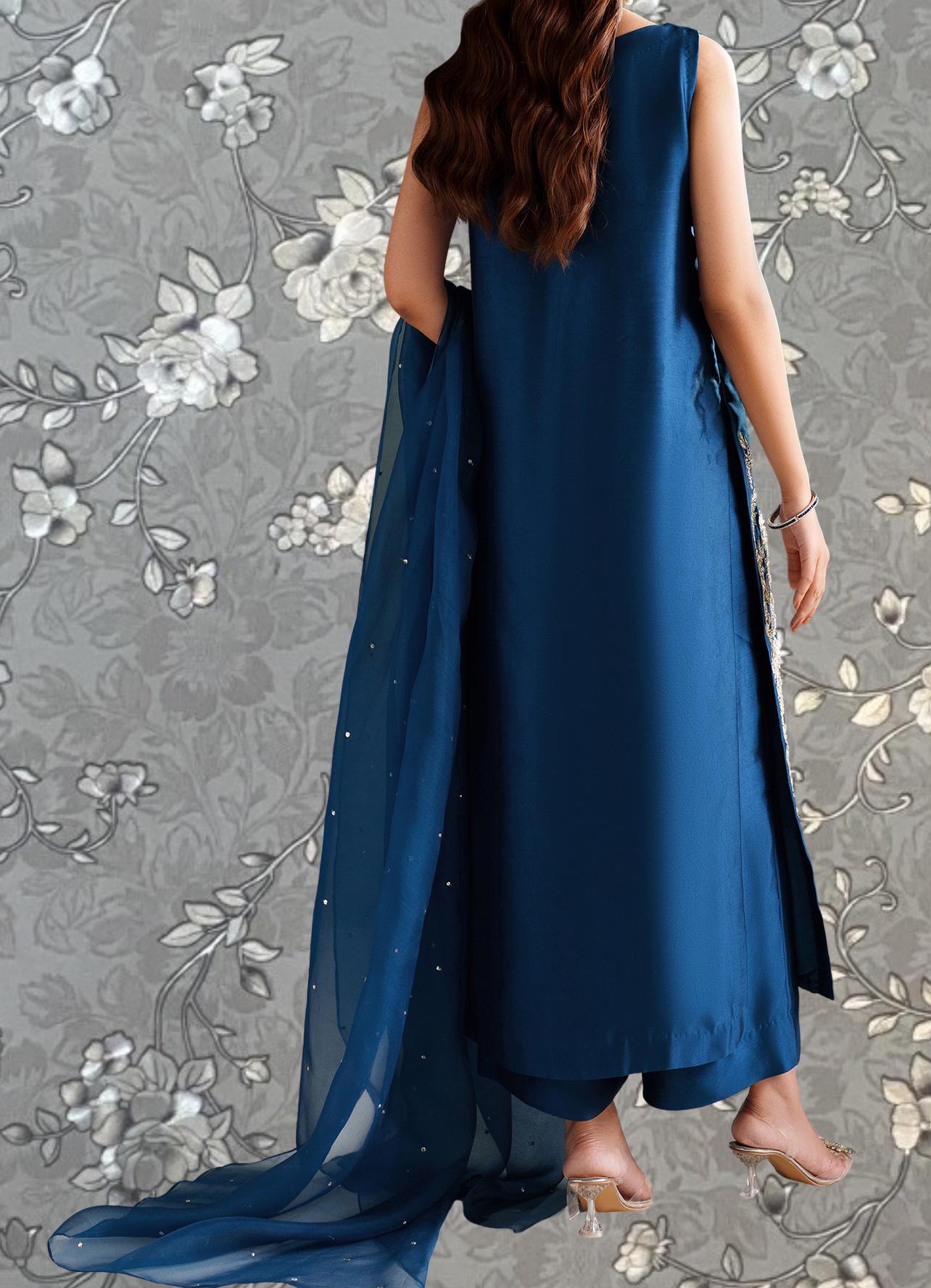 A woman wearing a long blue dress with a matching blue shawl and gold embellishments, standing against a gray floral background.