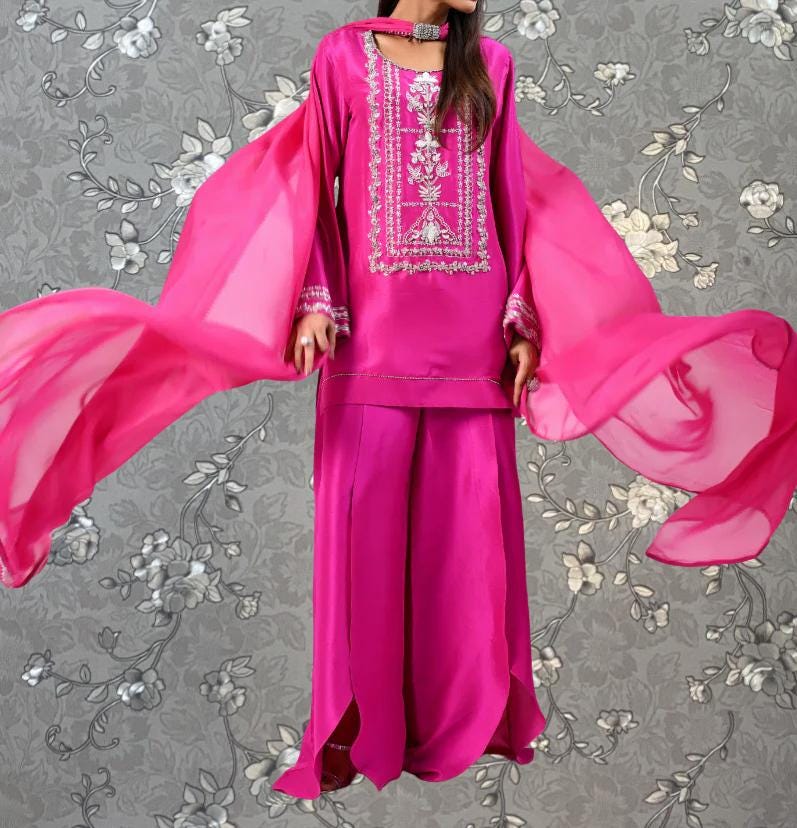 A woman wearing a bright pink dress with intricate embroidery and flowing sleeves stands against a gray floral background.