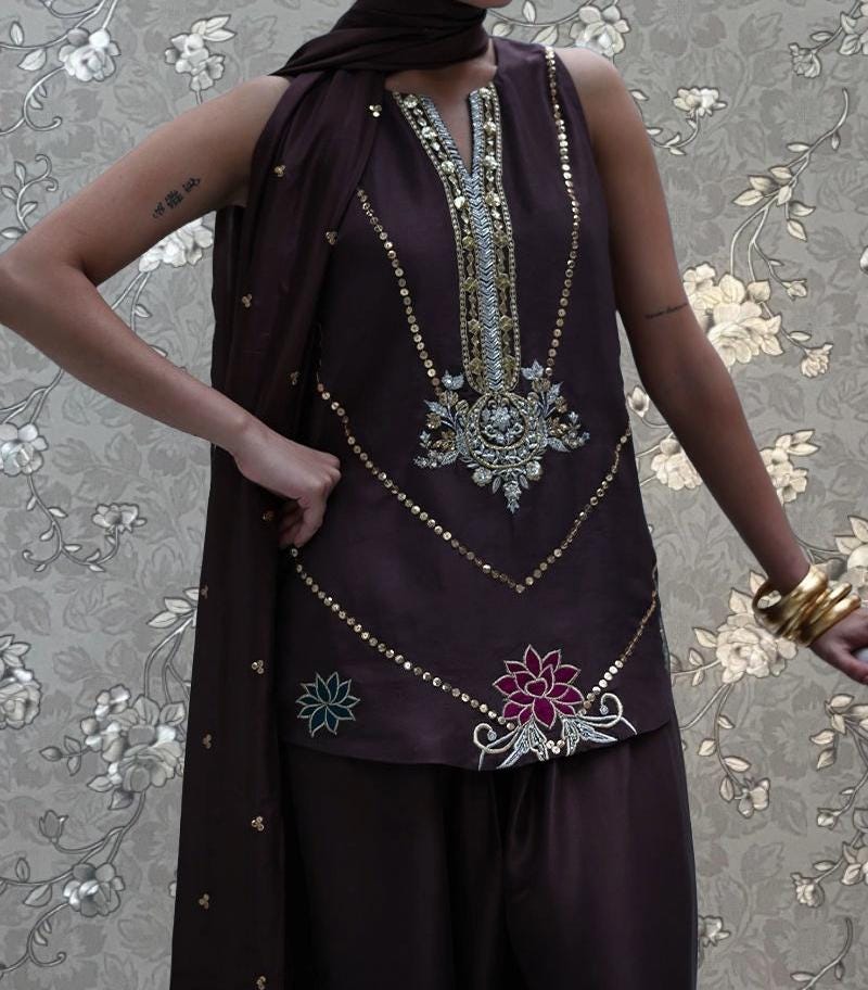A person wearing a purple dress with intricate embroidery and floral designs, posing against a floral patterned background.
