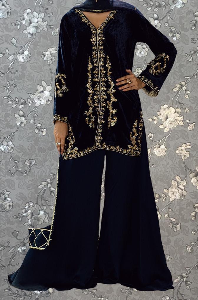 A person wearing a long, black velvet dress with gold embroidery and a matching black skirt, posing against a gray floral background.