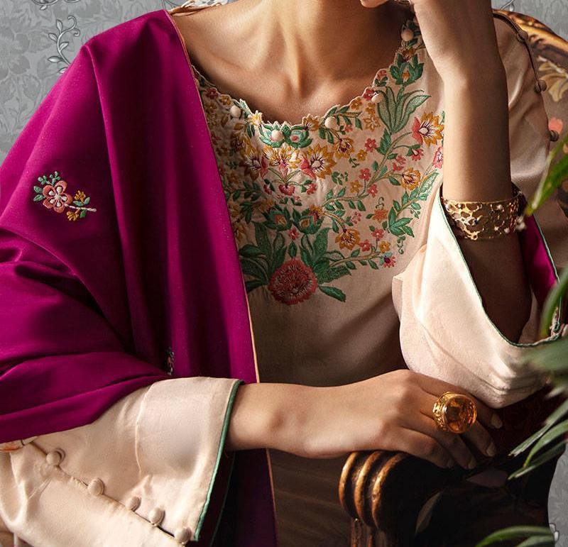 A close-up of a person wearing a pink shawl and a white blouse with a floral embroidered design, holding a gold ring on their finger.