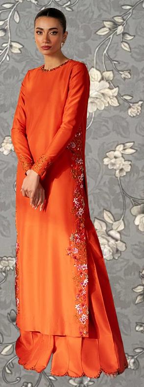 A woman wearing a long, orange dress with floral embroidery stands against a gray floral background.