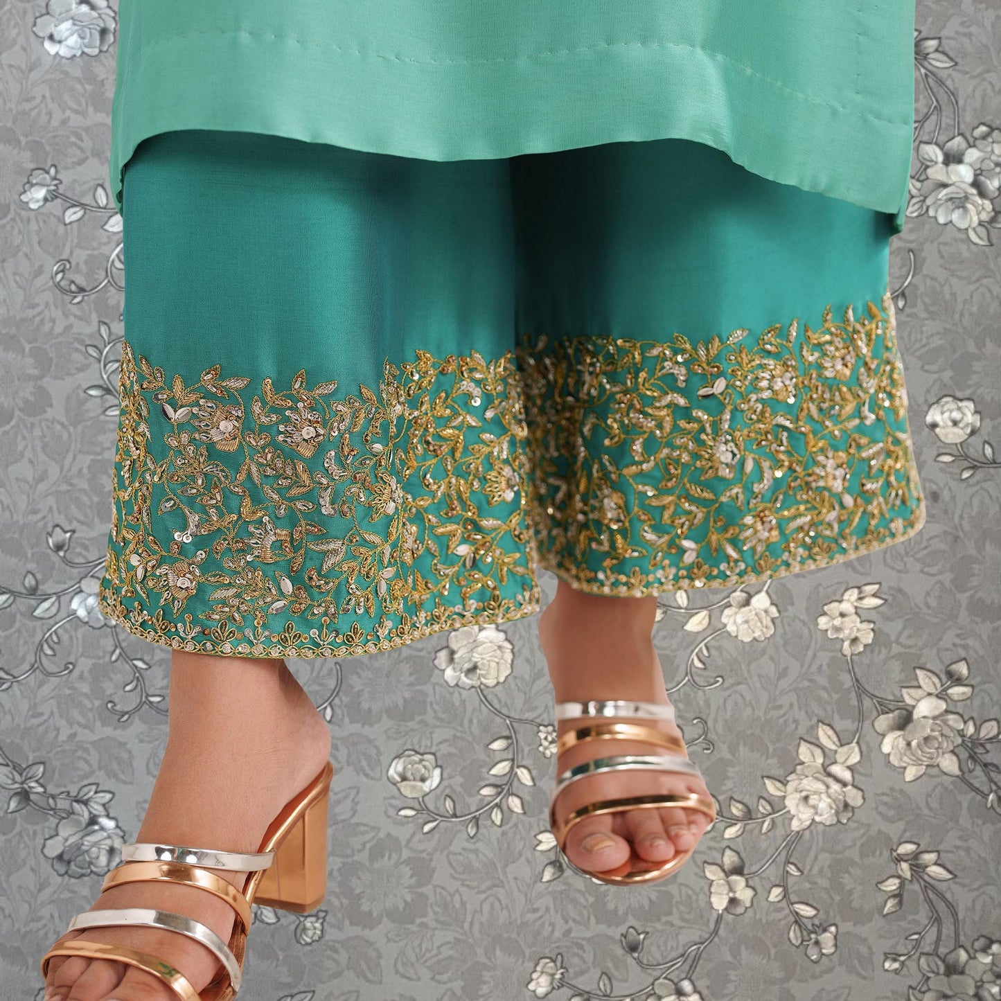 A close-up view of a person's legs wearing a green and gold embroidered dress, with gold heels visible.